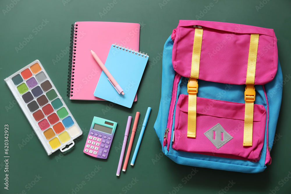 Colorful school backpack with notebooks, watercolors and calculator on green background