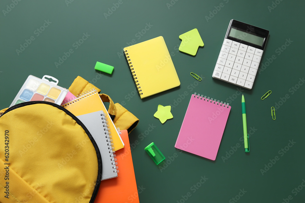 Yellow school backpack with notebooks, watercolors and calculator on green background