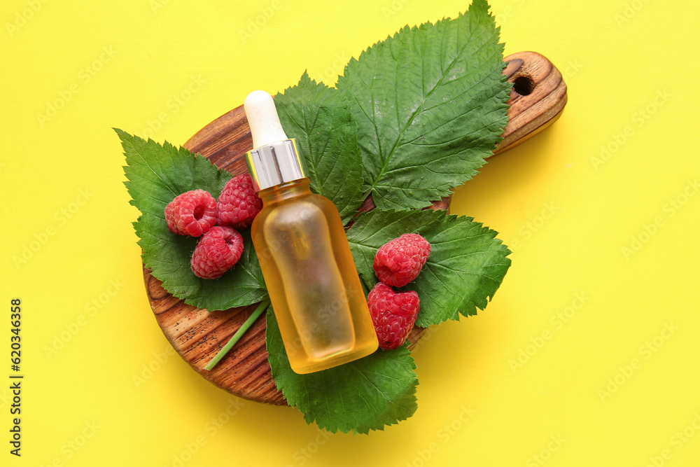 Wooden board with bottle of cosmetic raspberry oil on yellow background