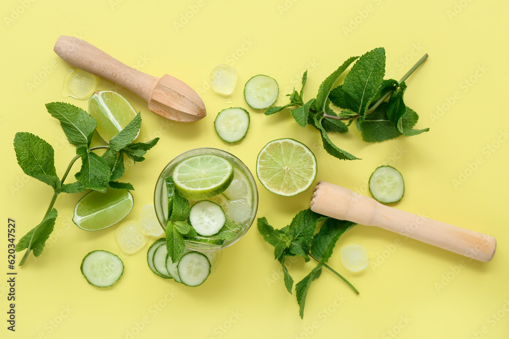 Glass of cucumber mojito with ingredients and juicers on yellow background