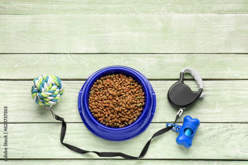 Bowl of dry pet food, leash, waste bags and toy on color wooden background