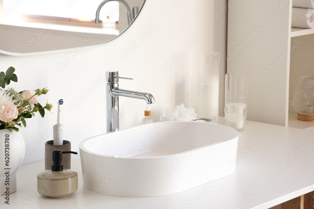 White sink with bath accessories and flowers on table in light bathroom, closeup