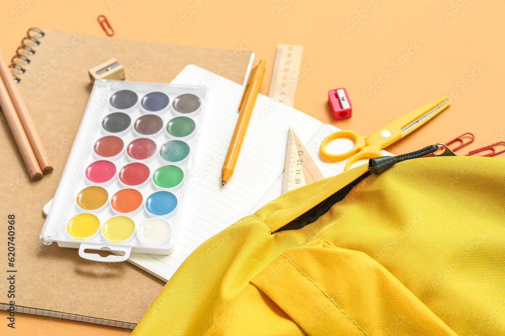 School backpack with paints and stationery on orange background, closeup