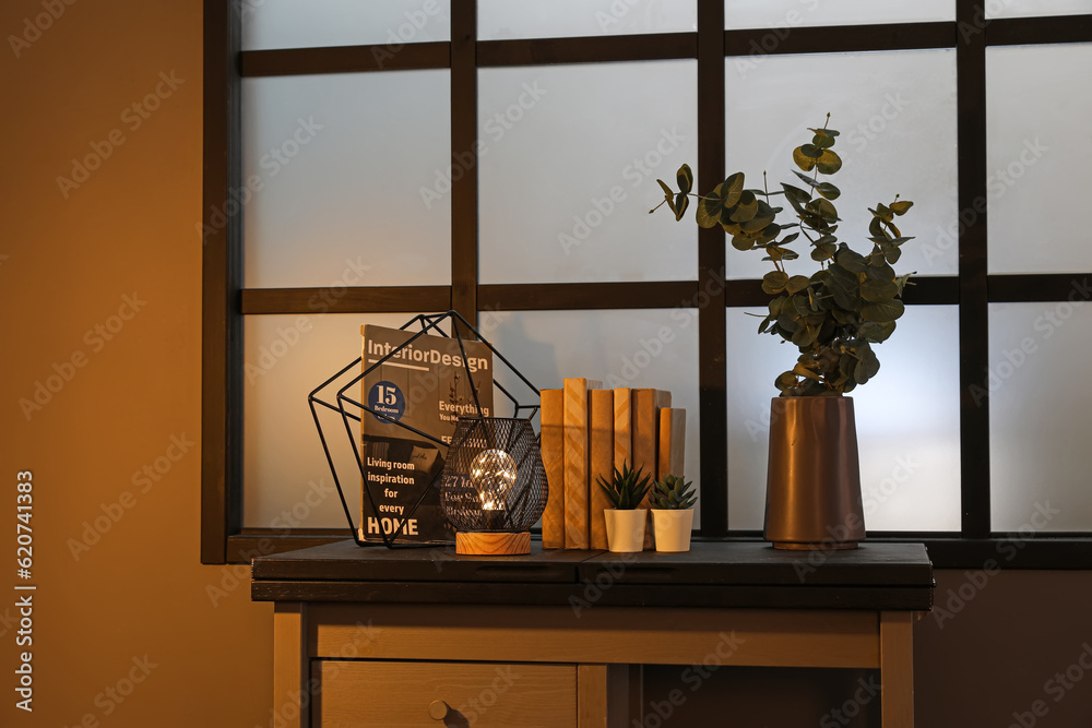 Table with glowing lamp, decor and houseplants in dark living room