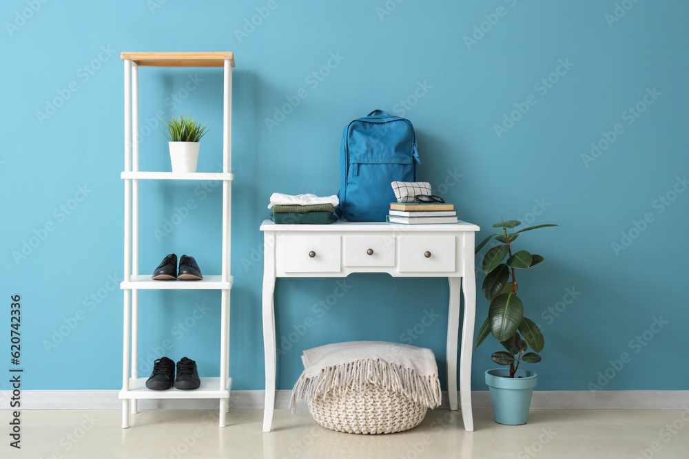 Table with stylish school uniform, backpack, shelving unit and pouf near blue wall in room