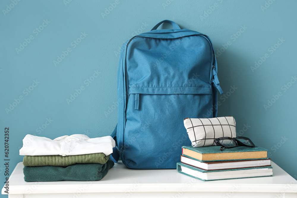 Table with stack of stylish school uniform, backpack, books, eyeglasses and pencil case near blue wa