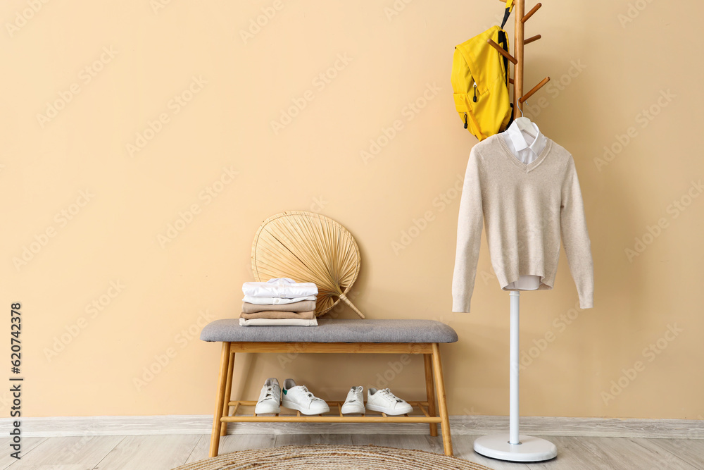 Stylish school cardigan with shirt, backpack hanging on rack, bench and sneakers near beige wall in 