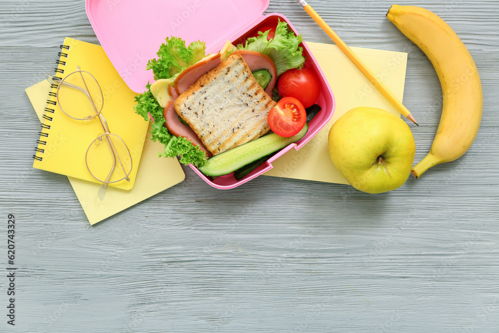 Stationery, eyeglasses and lunch box with tasty food on grey wooden background