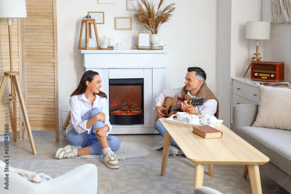 Young woman and her husband playing guitar near fireplace at home