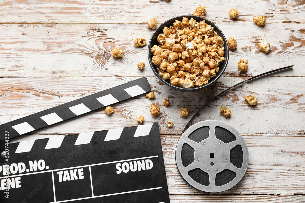 Bowl with tasty popcorn, clapperboard and film reel on white wooden background