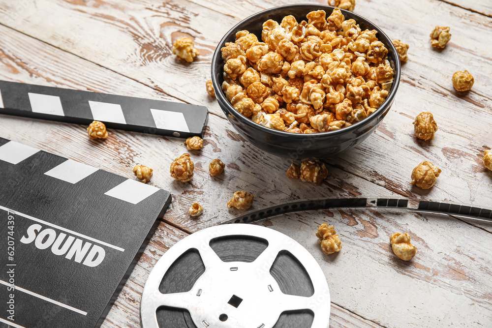 Bowl with tasty popcorn, clapperboard and film reel on white wooden background