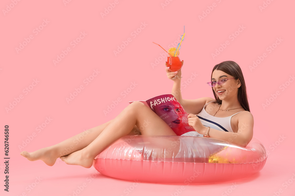 Young woman with cocktail and inflatable ring reading magazine on pink background