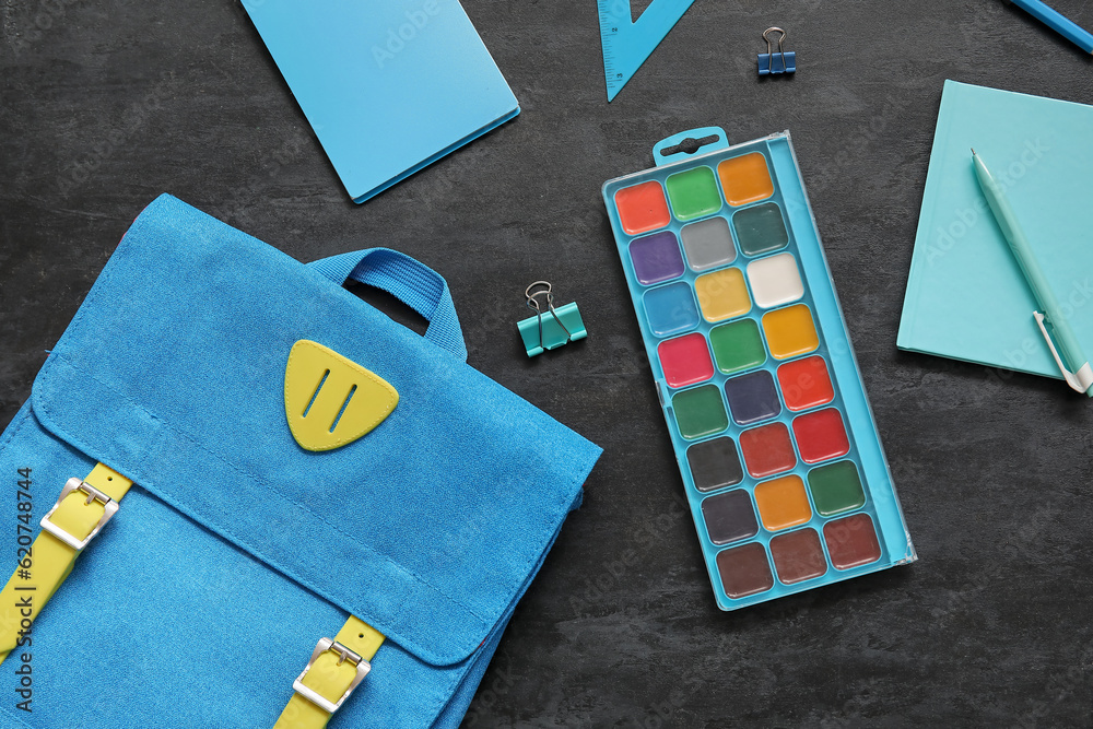 Blue school backpack with watercolors, notebooks and paper clips on black background