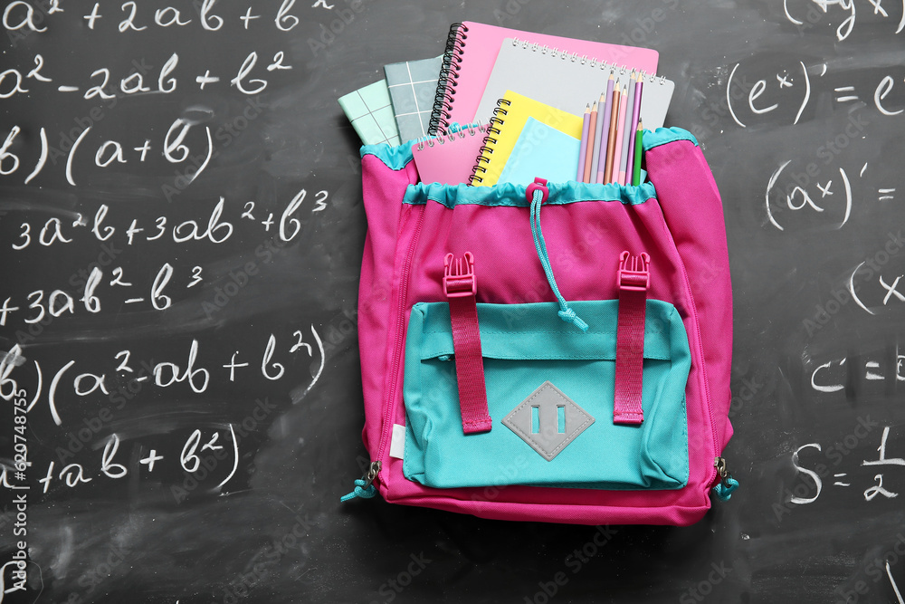 Colorful school backpack with notebooks and pencils on black chalkboard painted with formulas