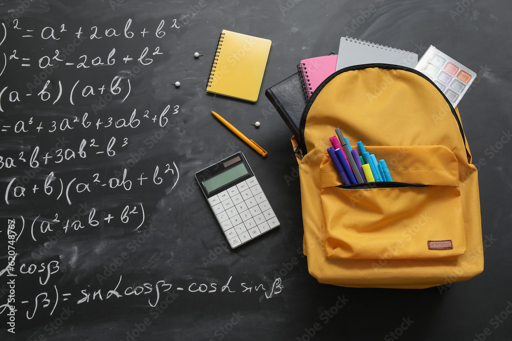 Yellow school backpack with notebooks, calculator and markers on black chalkboard painted with formu