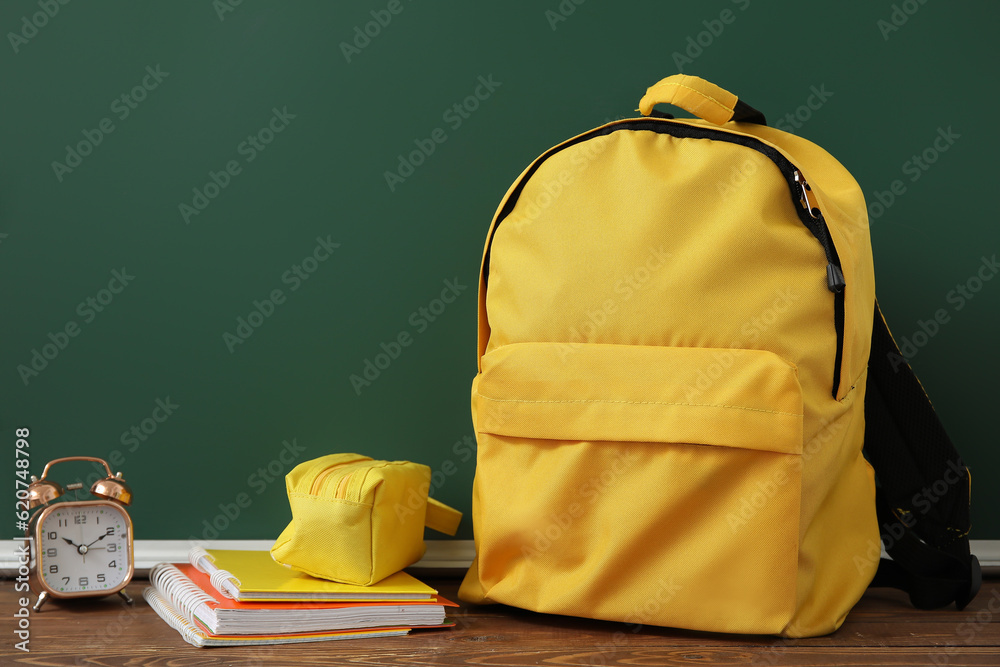 Yellow school backpack with alarm clock, pencil case and notebooks on wooden table near green chalkb