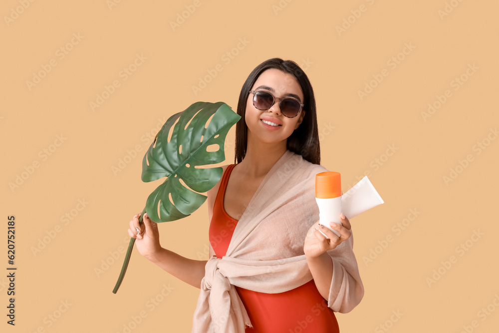 Beautiful woman with bottles of sunscreen cream and palm leaf on beige background