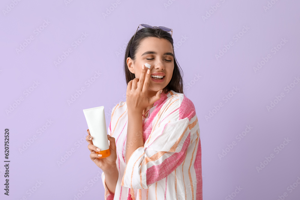 Beautiful young woman applying sunscreen cream on lilac background