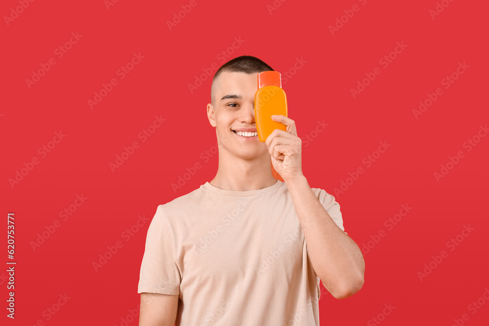 Happy young man with bottle of sunscreen cream on red background