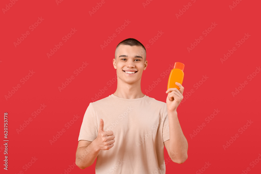 Happy young man with bottle of sunscreen cream showing thumb-up gesture on red background