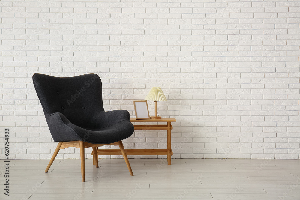 Stylish black armchair, wooden table and lamp near white brick wall