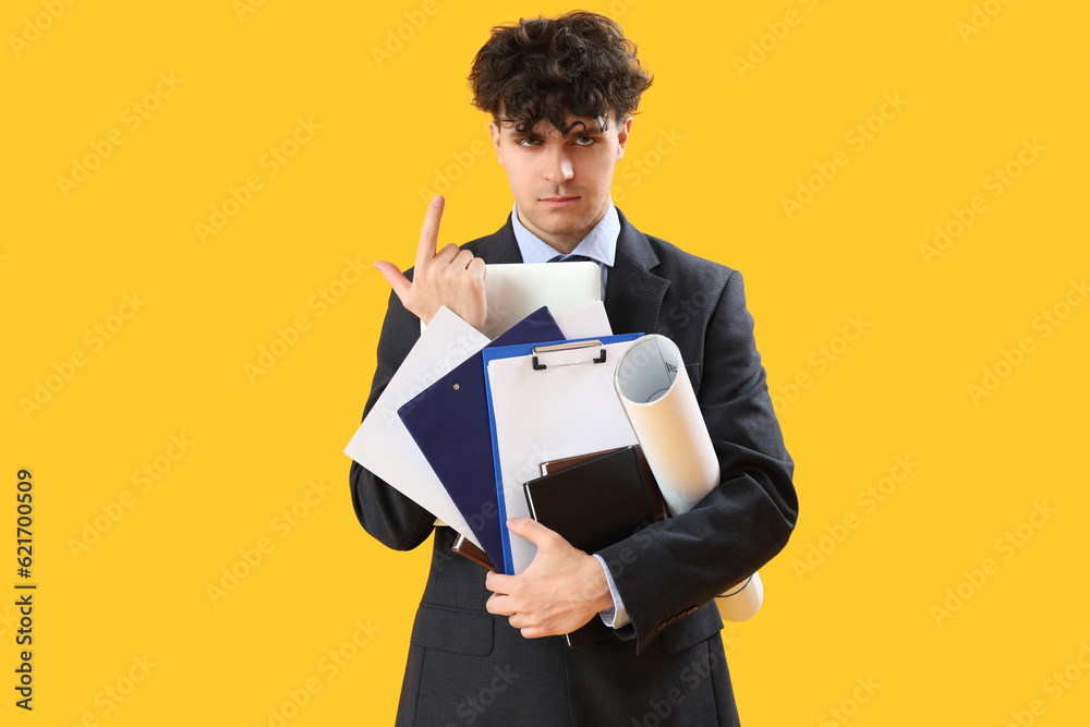 Young businessman with documents showing loser gesture on yellow background