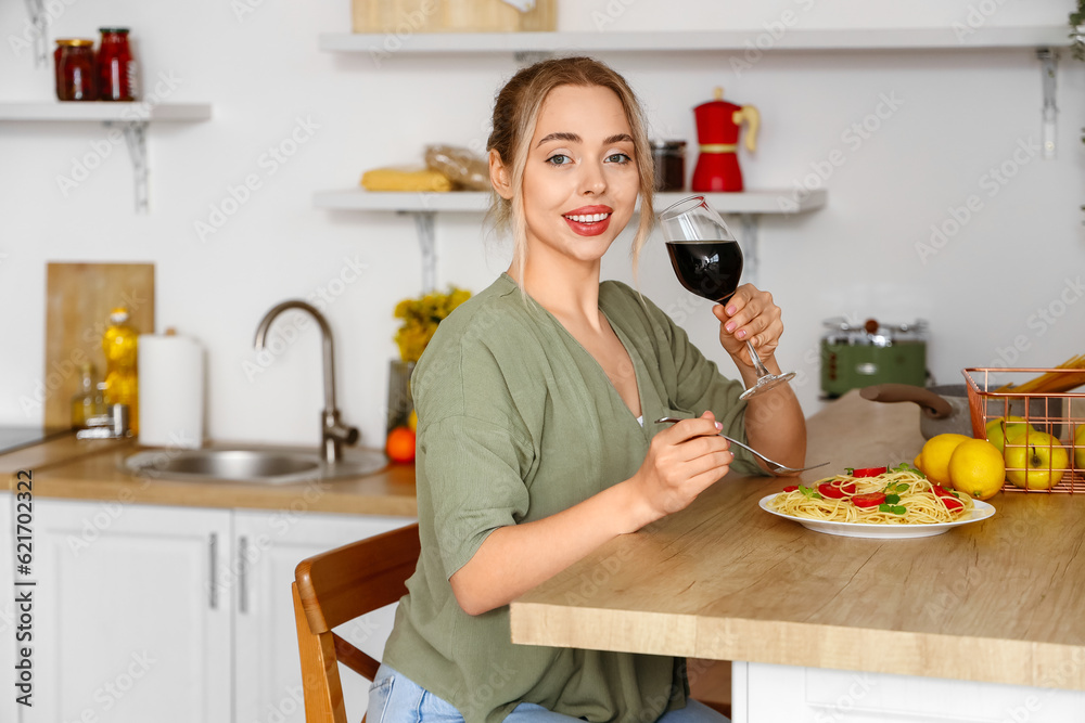 Young woman with glass of wine eating tasty pasta in kitchen