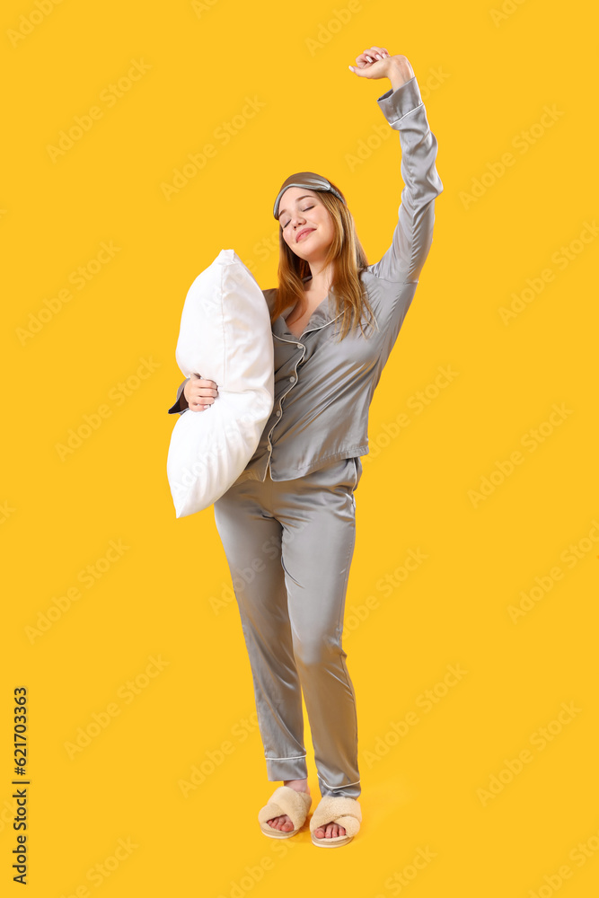 Young woman in pajamas with pillow on yellow background