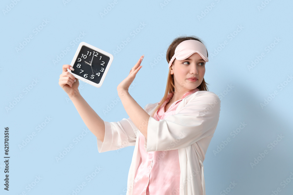 Young woman in pajamas rejecting clock on blue background