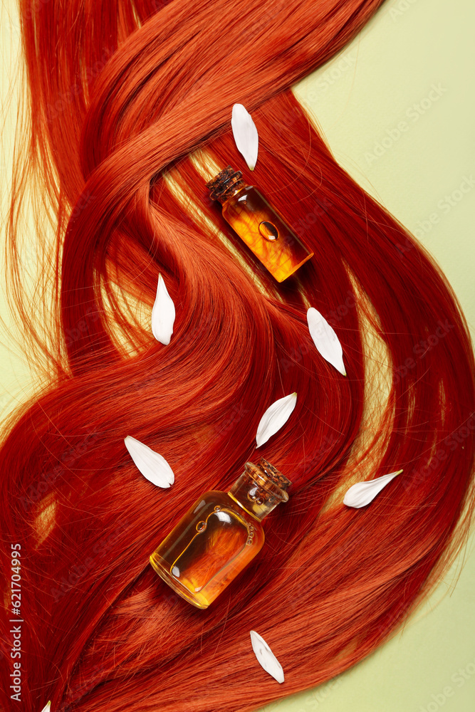 Composition with ginger hair strand, bottles of essential oil and flower petals on green background,