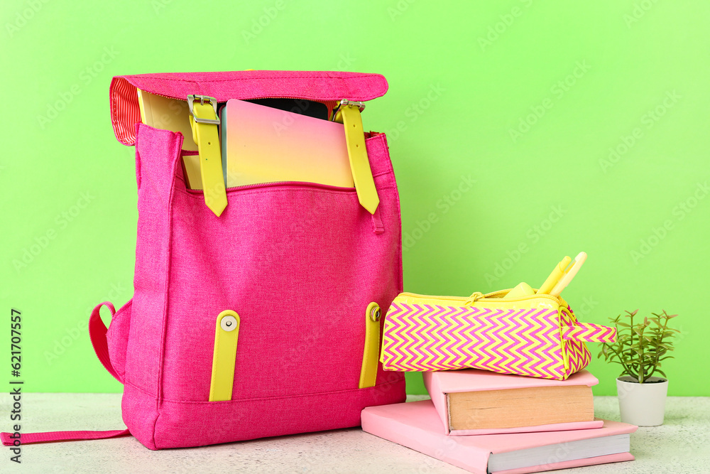 Pink school backpack with books, pencil case and houseplant on white wooden table near green wall