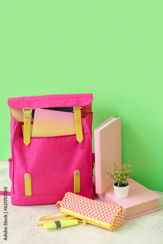 Pink school backpack with books, pencil case and houseplant on white wooden table near green wall