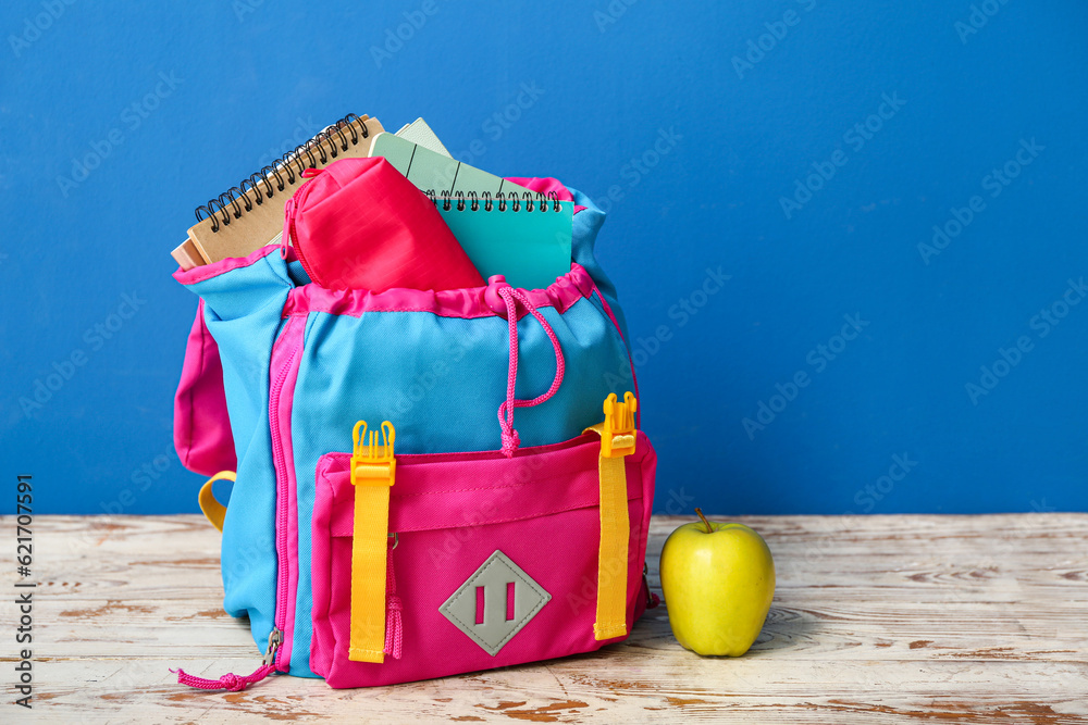 Color school backpack with notebooks, pencil case and apple on wooden table near blue wall