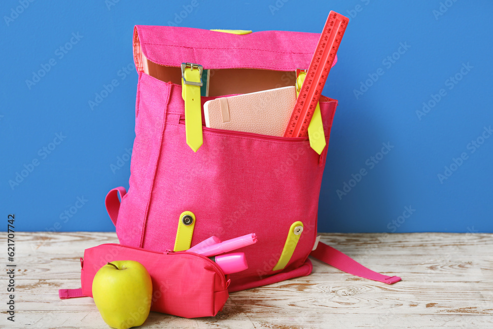 Pink school backpack with pencil case, markers and apple on wooden table near blue wall