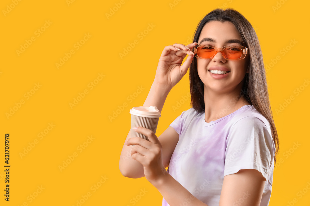 Beautiful young woman wearing stylish sunglasses and drinking coffee on yellow background