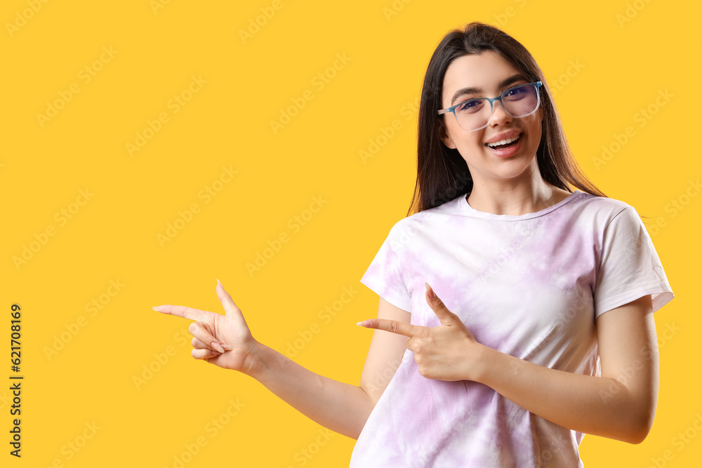 Pretty young woman wearing glasses and pointing at something on yellow background