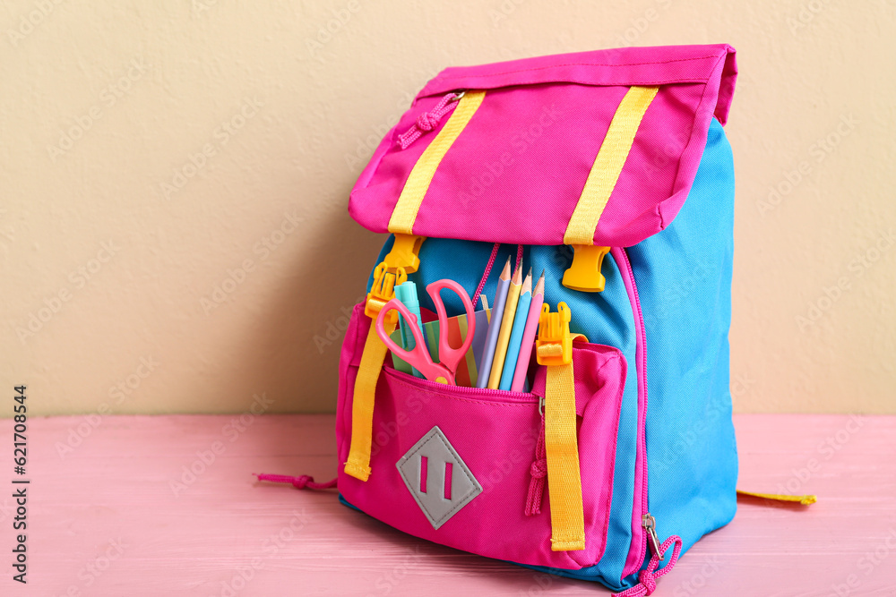 Color school backpack with scissors, pencils and notebook on wooden pink table near beige wall