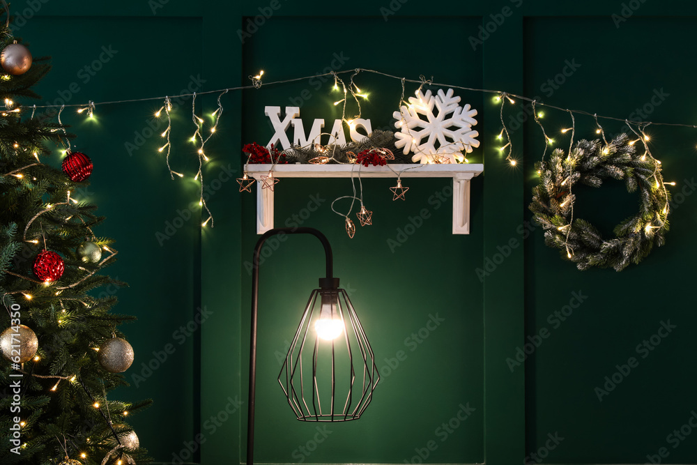 Glowing lamp, shelf with Christmas decor and wreath hanging on green wall in living room