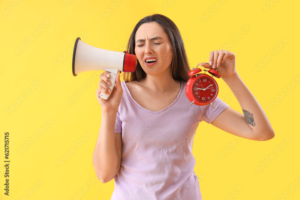 Stressed young woman with alarm clock shouting into megaphone on yellow background. Deadline concept