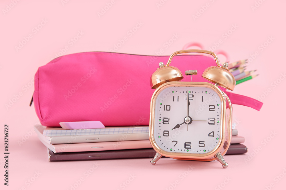 Alarm clock with notebooks and pencil case on pink background