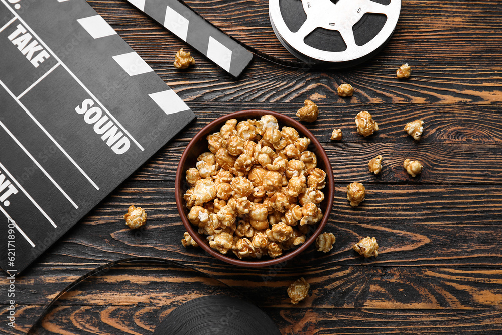 Bowl with tasty popcorn, clapperboard and film reel on wooden background