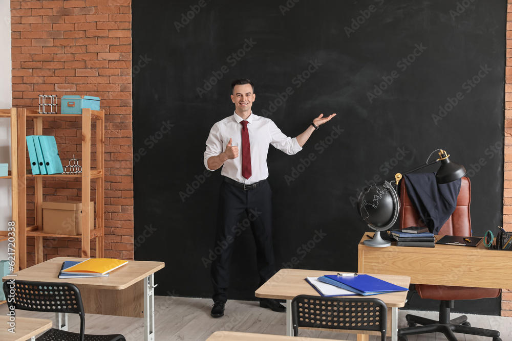 Male teacher showing thumb-up near blackboard in classroom