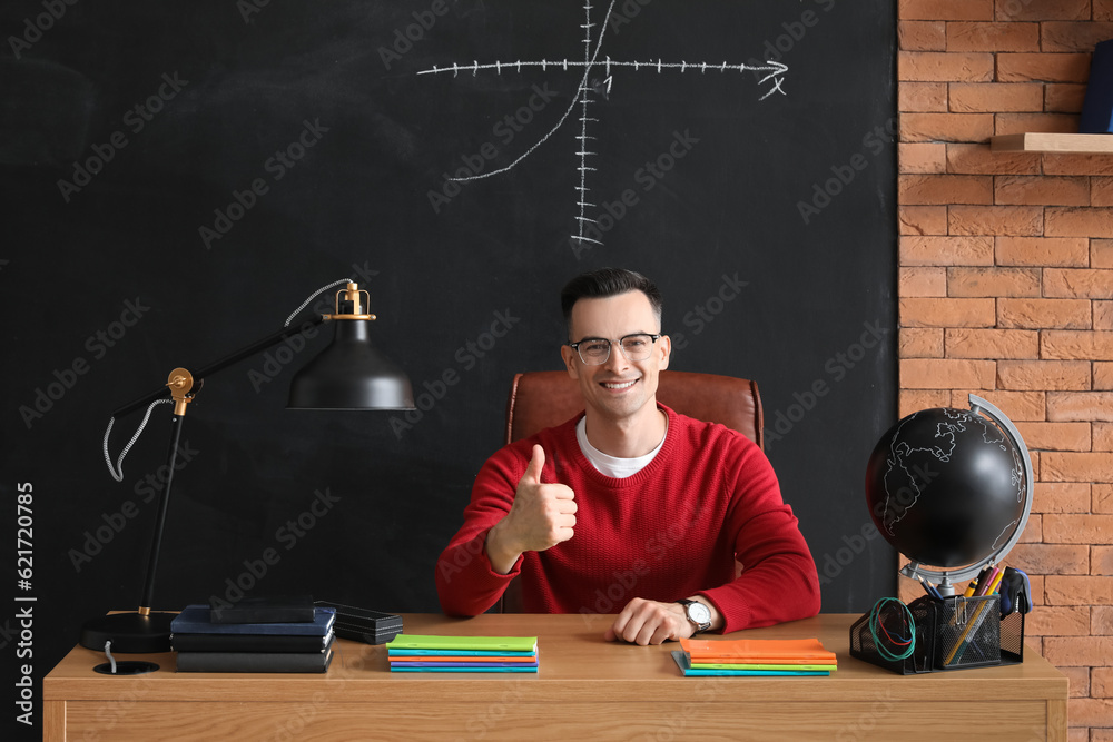 Male teacher showing thumb-up at table in classroom