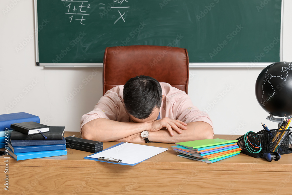 Tired male teacher sitting at table in classroom