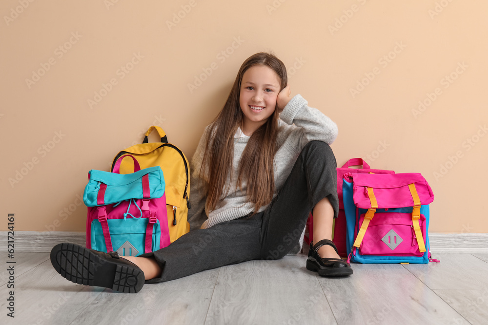 Little schoolgirl with backpacks sitting near beige wall