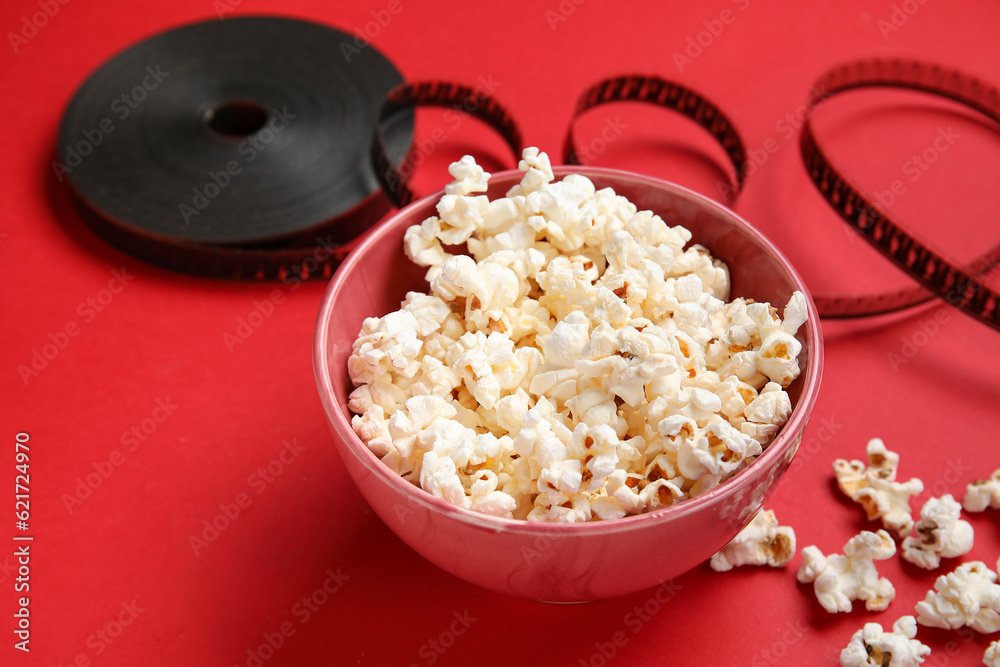 Bowl with tasty popcorn and film reel on red background