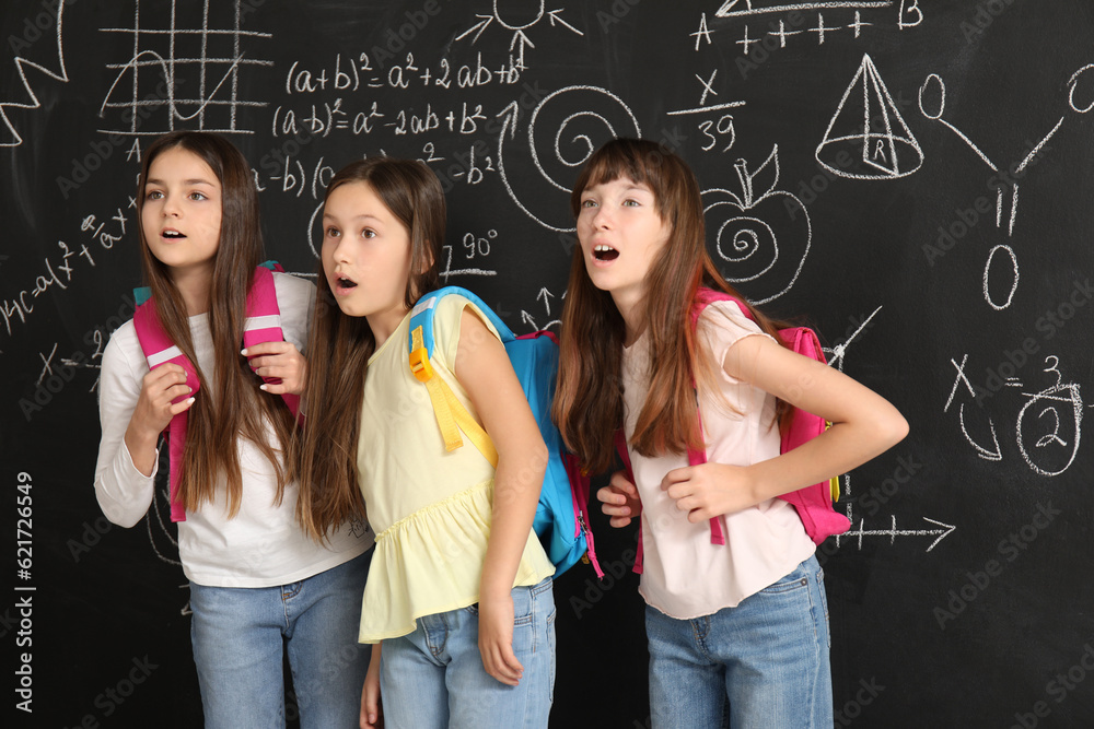 Shocked little girls with backpacks near blackboard