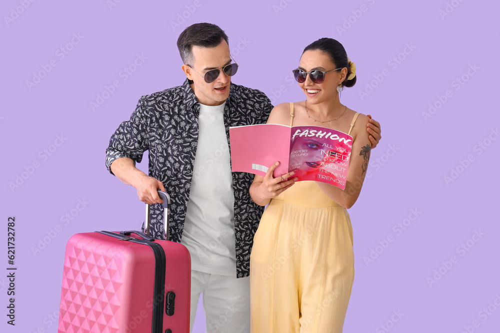 Young couple with magazine and suitcase on lilac background