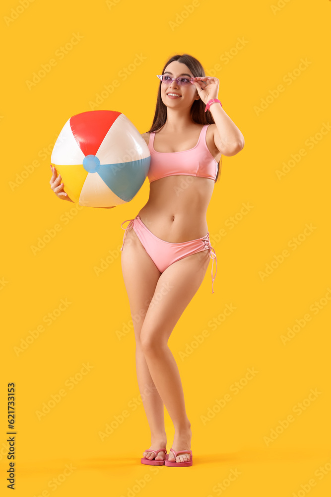 Young woman in swimsuit with beach ball on yellow background