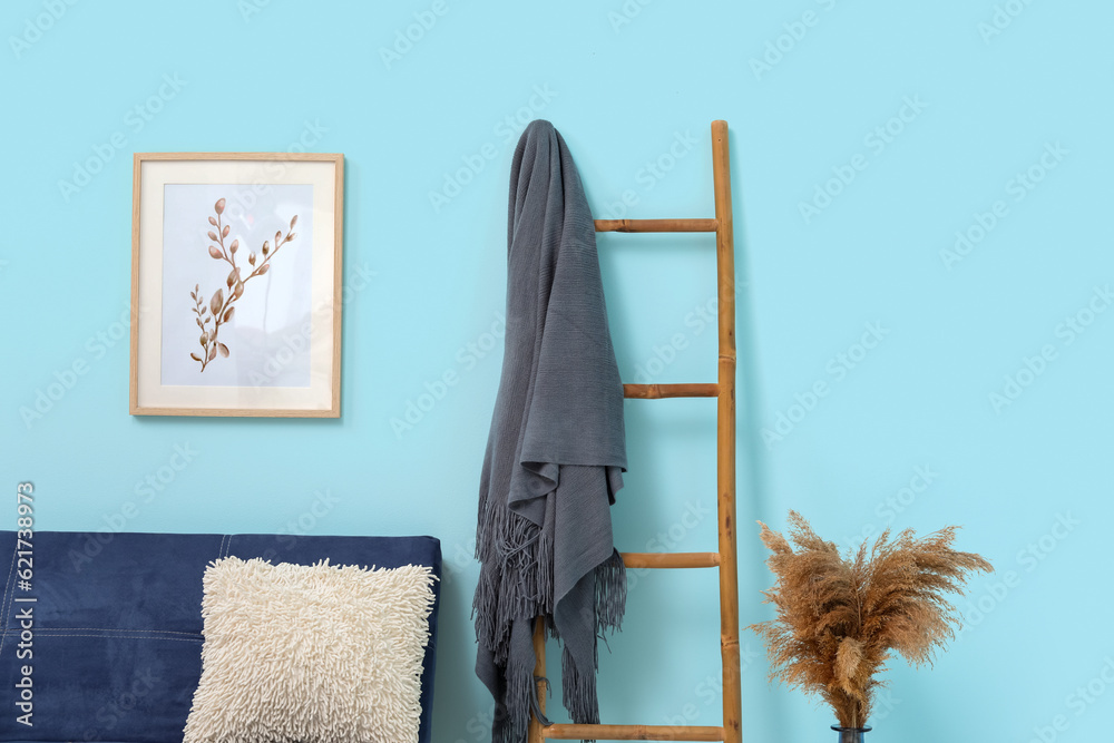 Wooden ladder with blanket, sofa, pampas grass and picture hanging on blue wall in room